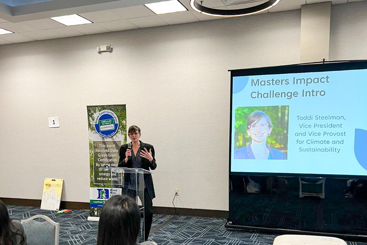 Person standing at a lectern and talking into a microphone. Next to her is a screen displaying a slide that reads Masters Impact Challenge Intro, Toddi Steelman, vice president and vice provost for climate and sustainability.