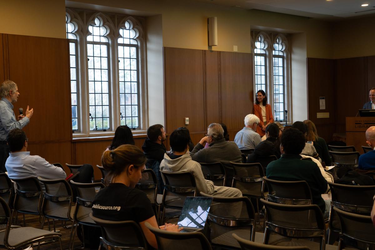 Among a seated audience, a person stands with a microphone.