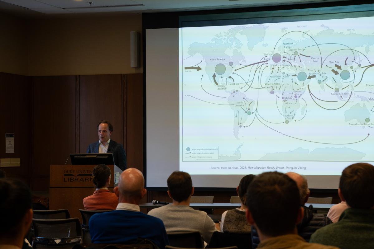 Person standing at a lectern and talking to a seated audience. Next to the person is a screen displaying a world map.