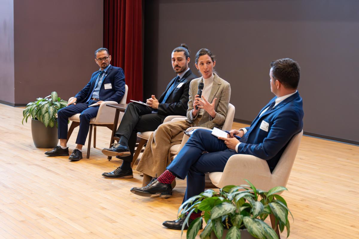 Four people sitting on a stage. One is speaking into a microphone.