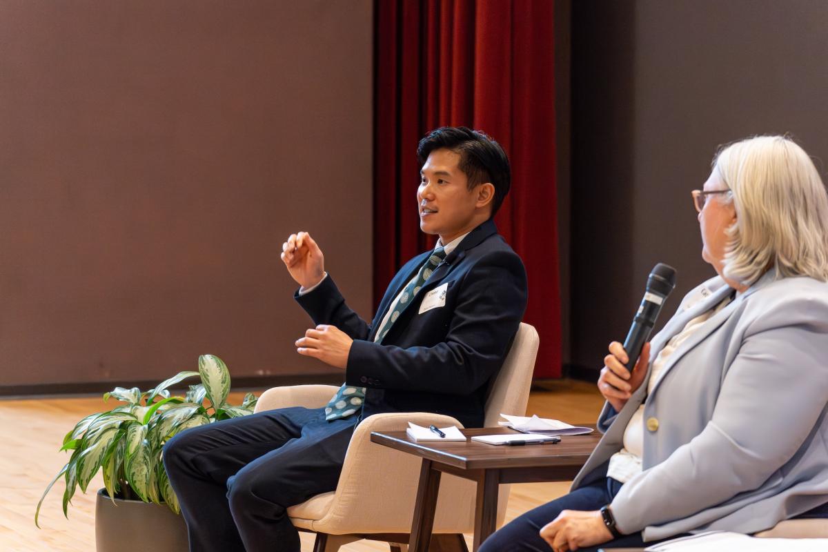 Two people speaking and sitting on a stage. One is holding a microphone.