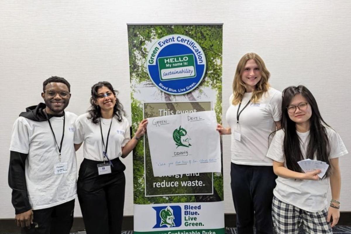 Four students standing together by a banner that says Bleed Blue. Live Green. I support Sustainable Duke. Two of the students are holding a sign that says Green Devil Dining.