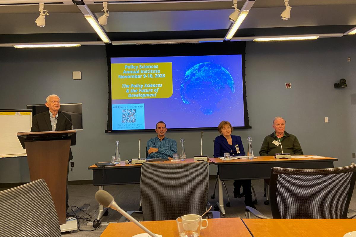 Three panelists sitting at a table. A moderator stands next to them at a lectern.