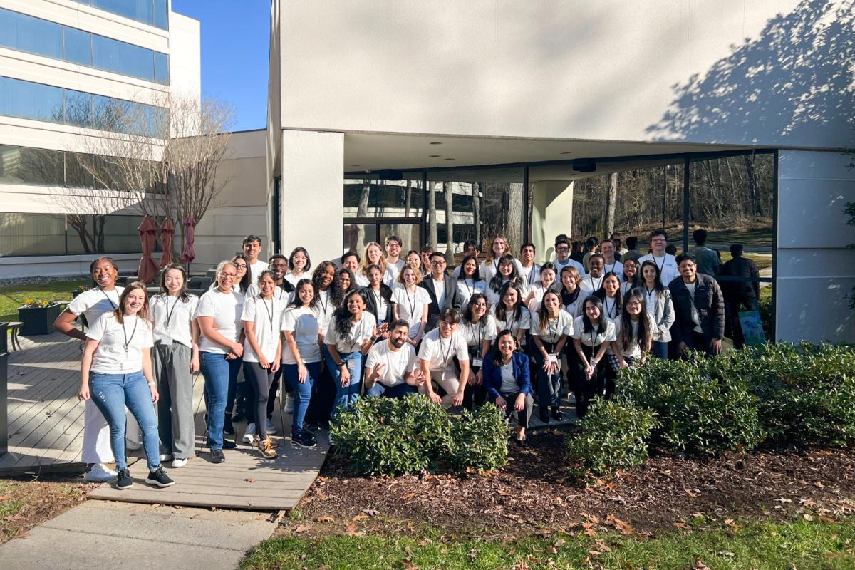 Several people gathered together and smiling. They are standing in front of a building and most are wearing white t-shirts.