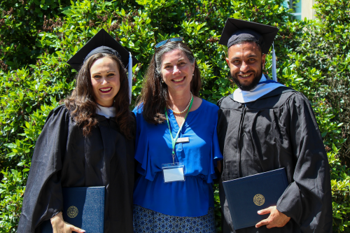 A male and a female student dressed in graduation caps and gowns standing with staff member